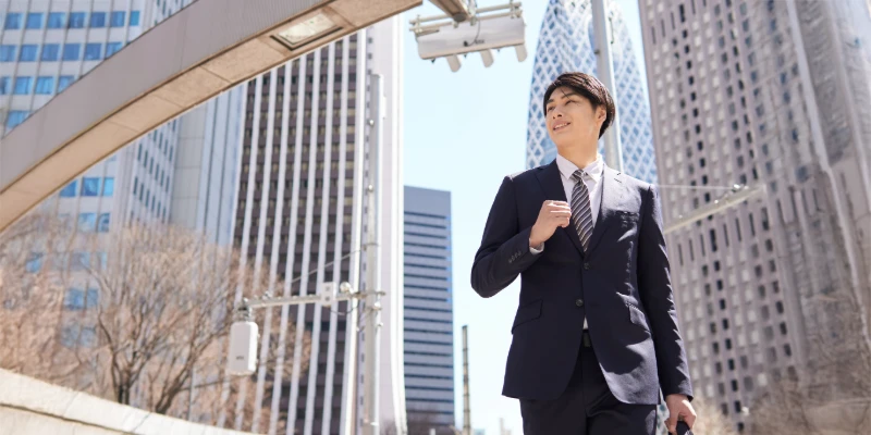 スーツを着た日本人ビジネスマンが新宿の高層ビル街を歩く様子。キャリア成功の象徴。
