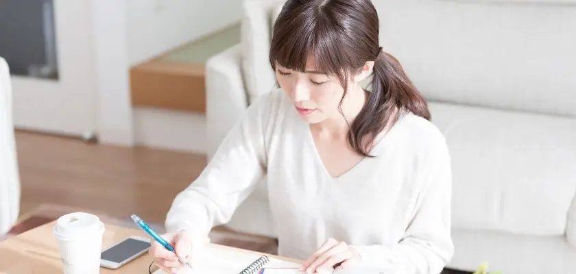 ノートに書き込む女性 白いトップスを着た女性が自宅のテーブルでノートに書き込んでいる様子。スマートフォンとマグカップが近くに置かれている。
