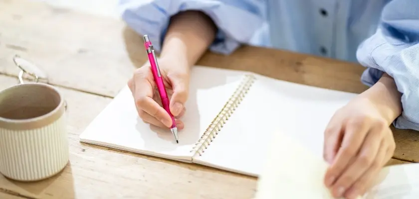 ノートにボールペンで書き込む人の手元とコーヒーカップ