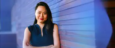 Image of a confident woman in a sleeveless navy top standing with arms crossed, smiling slightly while leaning against a modern wooden-paneled wall in a brightly lit setting.