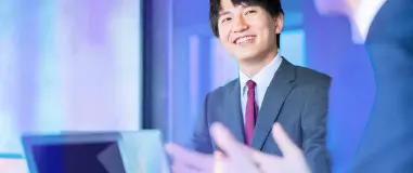 Business meeting scene showing a smiling man in a suit and red tie, seated at a table with a laptop, engaging with a colleague in a bright, modern office environment with a cool blue-toned color scheme.