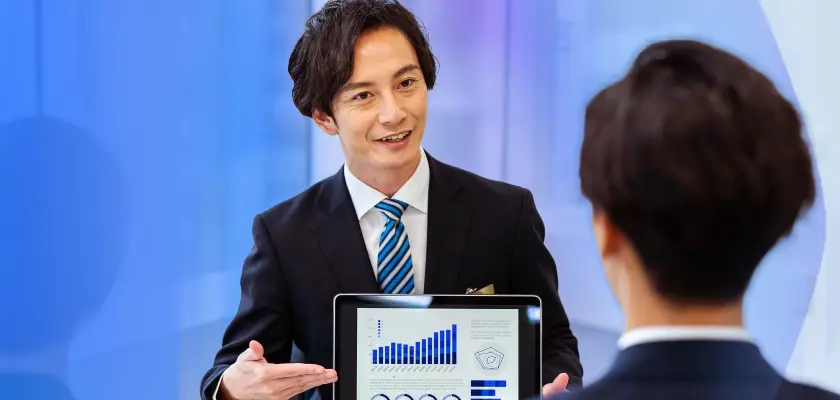 スーツ姿の男性がタブレットの画面を示しながら説明する様子、グラフが表示された画面