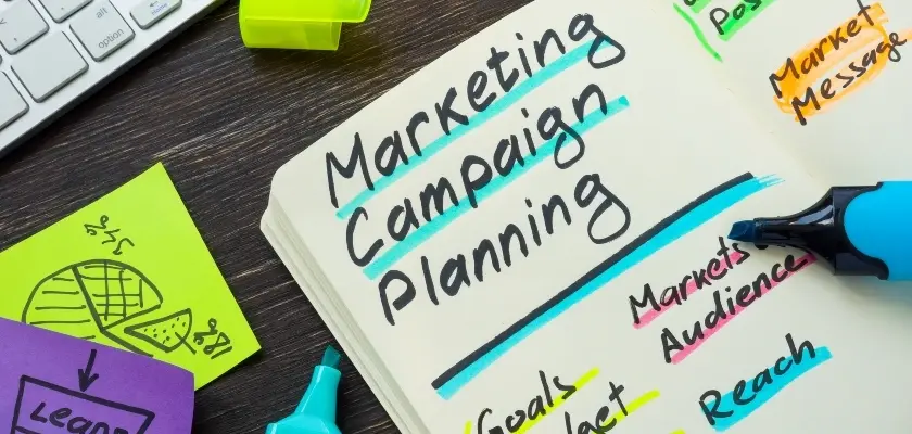 マーケティングキャンペーン企画ノート 「Marketing Campaign Planning」と書かれたノート。「Market Audience」「Reach」「Market Message」などの関連用語がハイライトされており、カラフルな付箋とキーボードが周りに配置されている。